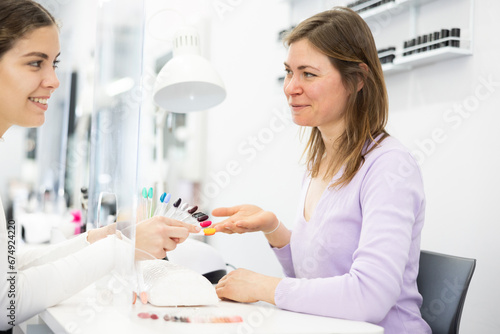 Positive woman nail technician offering to young female client to choose color of nail polishes in beauty salon..
