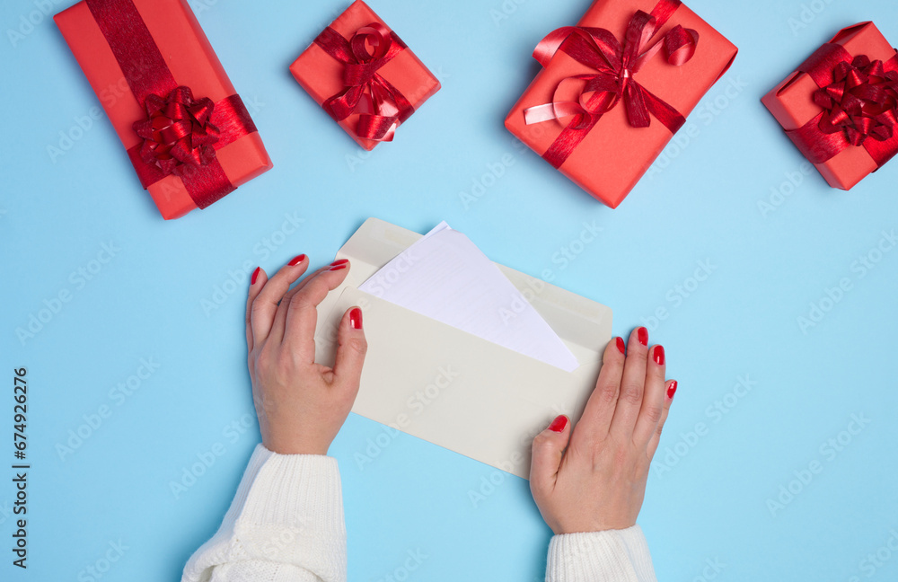 Female hands holding a white paper envelope on a blue background, around a box with gifts Female hands holding a white paper envelope on a blue background, around a box with gifts