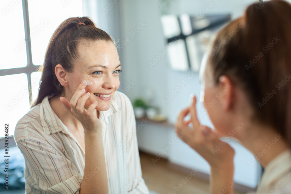 Beautiful young woman applying facial cream for healthy skin with mirror Beautiful young woman applying facial cream for healthy skin with mirror