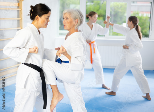 Elderly woman and young female karateka train karate kicks in group in studio