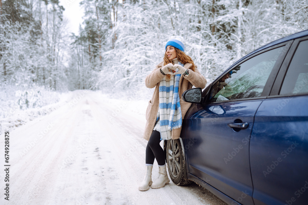 Beautiful young woman in a blue hat and scarf stands near a car on a snowy forest road. A traveler poses near a car in nature. Concept of car travel, vacation.