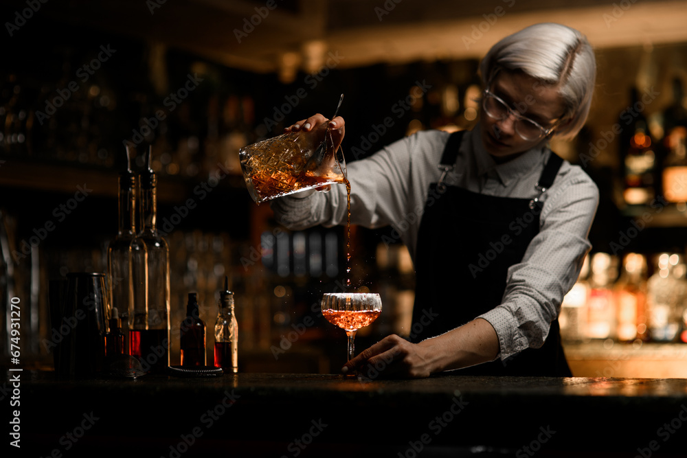 Female bartender pours an iced cocktail from a mixing glass into a cocktail glass, splashes fly to the sides