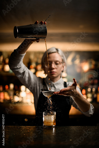 Female bartender pours a cocktail through a strainer from a metal glass into a glass with a piece of ice, splashes fly to the sides