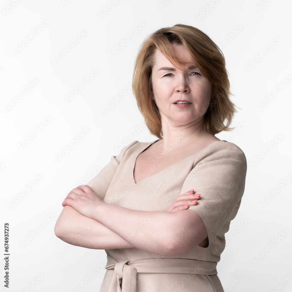 Serious woman 49 year old in beige dress looking at camera. Arms crossed on chest. White background