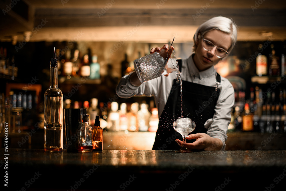 Female bartender prepares a cocktail with ice and a transparent drink in a cocktail glass on a stem, splashes fly