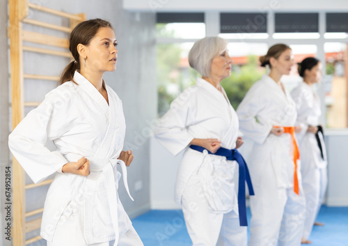 Concentrated young girl in white kimono practicing punches in gym during martial arts workout with female group. Shadow fight  combat sports training concept