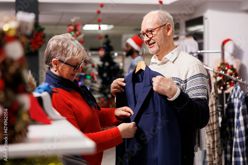 Elderly couple looking at blazers in retail store, searching for formal clothing to wear on christmas dinner celebration. Customers checking fabric of suit jacket for elegant outfit, festive decor. photo