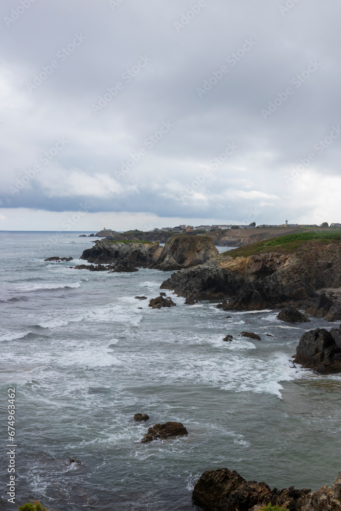 a rocky coastline with a cloudy sky, rough waters, and a small lighthouse in the background