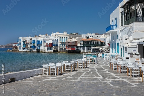 Row of wooden tables and chairs are situated along a paved stone walkway on the shore of Mykonos