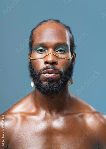 Black gay stands isolated on blue background and stares into camera with serious expression on face. Close-up portrait of handsome adult transgender with blue eyeshadows and golden accessory on face