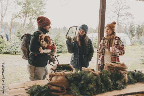 Friends with their dog having fun during Christmas tree harvesting at Eco Pine and Fir tree Farm. Casually dressed Happy family with Cavalier King Charles Spaniel hanging out outdoors, Holiday season. #675025655