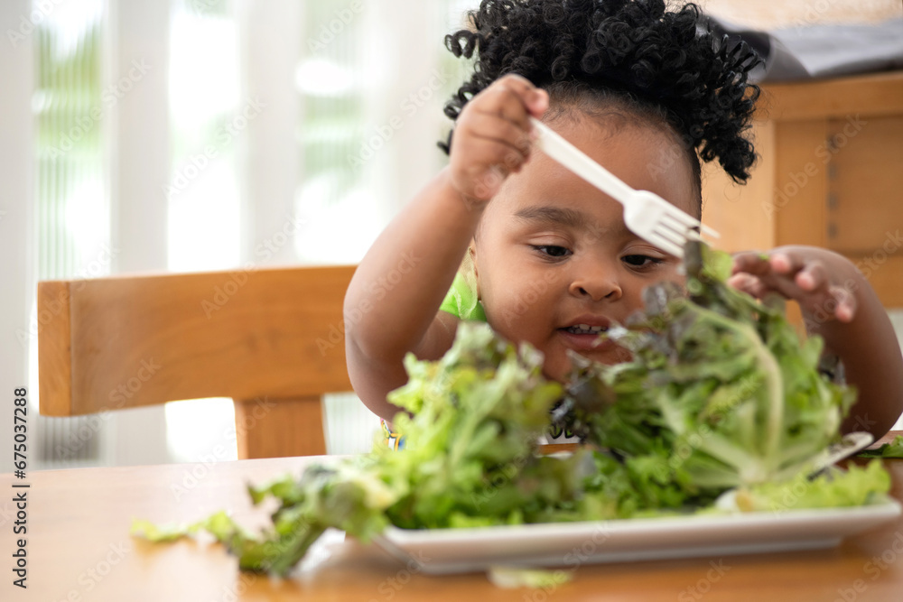 Cute little African girl likes to eat green vegetables, use her hands to pick up lettuce and put it in her mouth - obrazy, fototapety, plakaty 
