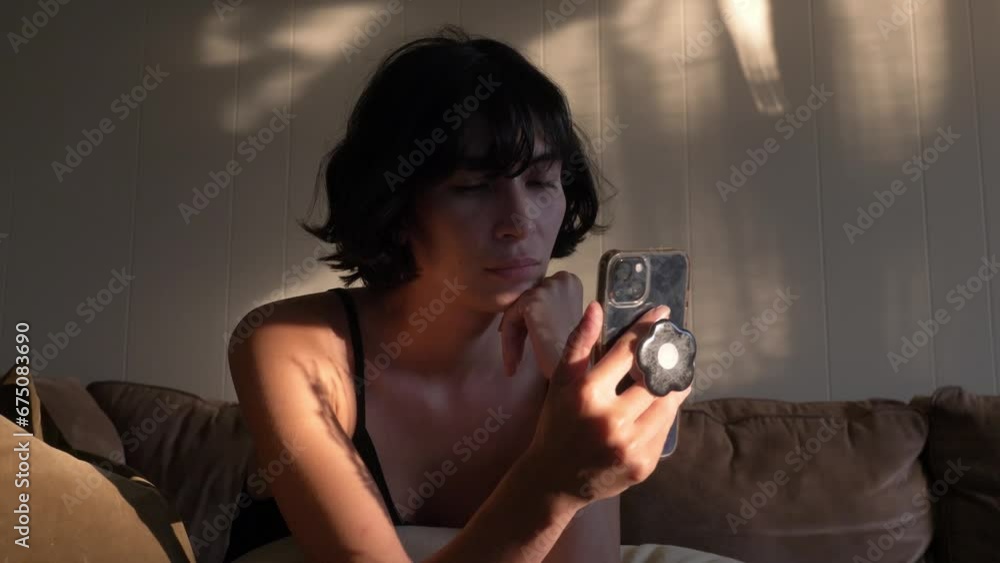 Girl illuminated by sunlight sitting on sofa while looking at smartphone. Handheld
