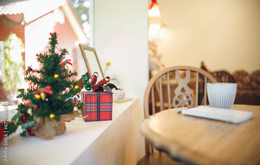 Decorate christmas event on counter by the window with red gift box and blurred small chrismas tree. On the side was a coffee cup and a notebook on a wooden table