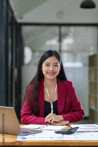 Confident Asian businesswoman smiling at camera while sitting at desk going through company financial business documents.