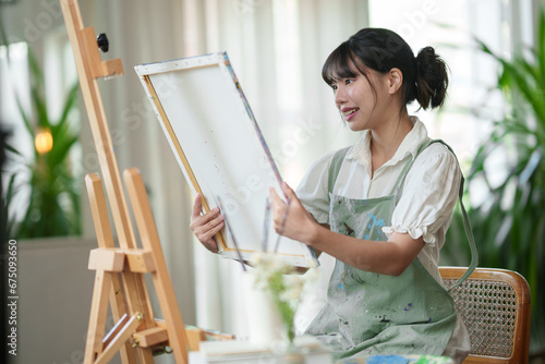 A female Asian artist who designs watercolor paintings with a paint brush sits holding a picture and happily admires her work. Painter's concept of using imagination on canvas. Hobbies in the studio.