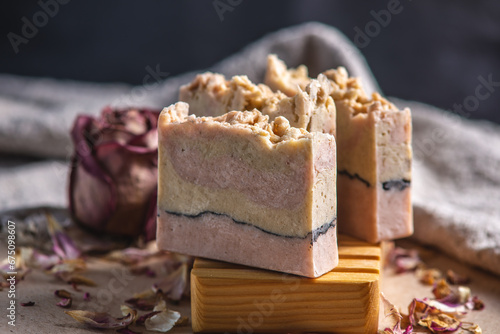 Delicate cute pieces of natural soap on a wooden soap dish on a background of fabric and pink flowers and petals nearby. Beautiful postcard