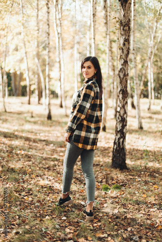 A beautiful girl in jeans and a plaid shirt walks in a birch grove. Portraits of a brunette girl in nature, in the autumn forest