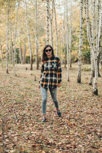 A beautiful girl in jeans and a plaid shirt walks in a birch grove. Portraits of a brunette girl in nature, in the autumn forest © Liudmila
