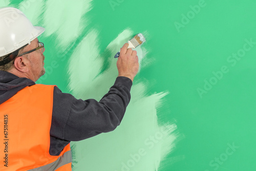 A painter in overalls and a helmet paints a wall with a brush. Copy space. green background