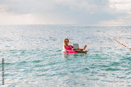 Woman works on laptop in sea. Freelancer, blond woman in sunglases floating on an inflatable big pink donut with a laptop in the sea at sunset. People summer vacation rest lifestyle concept. © svetograph