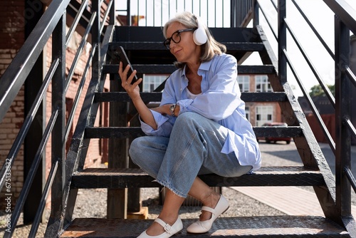 pretty fashionable middle-aged woman with gray hair dressed in summer style enjoys summer and listens to music with headphones