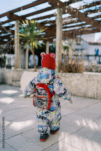 Small child with a backpack walks across the tiles in the yard to the pergola in the garden. Back view
