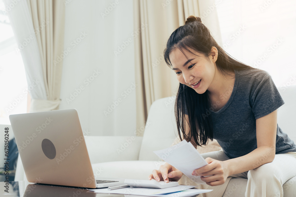 Young asian woman sits in the living room using a calculator to calculate family income and expenses and writes it down in his notebook or laptop at home.
