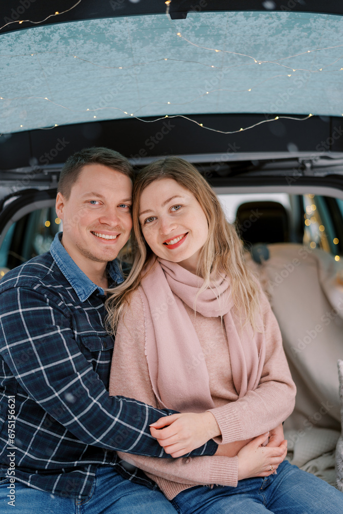 Hugging smiling couple sitting in car trunk on bedspreads