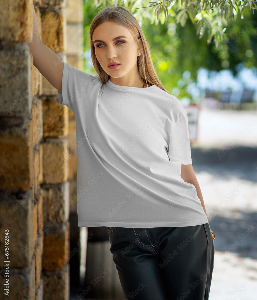 Mockup of a white T-shirt on a girl, leaning on a brick wall, on the background of trees, front view.