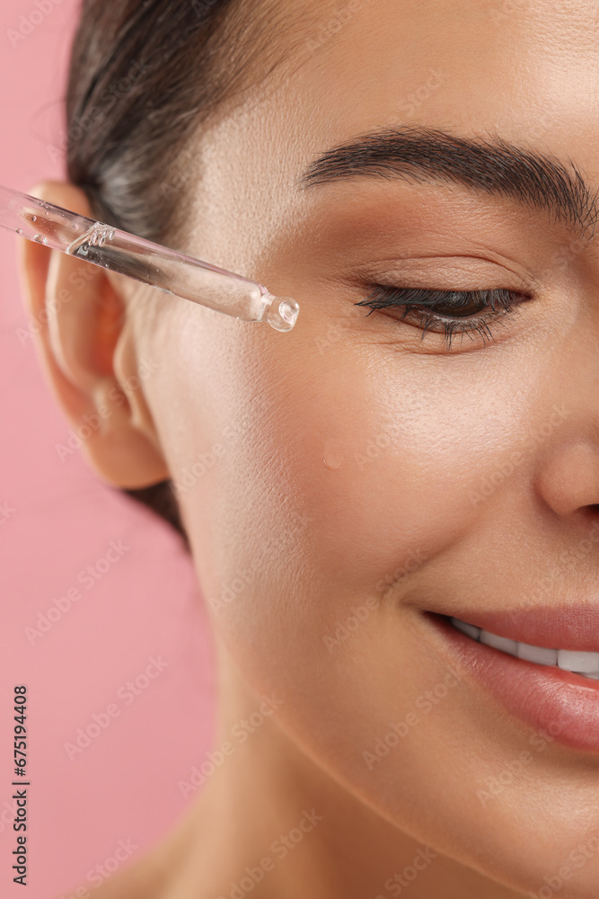 Beautiful young woman applying serum onto her face on pink background, closeup