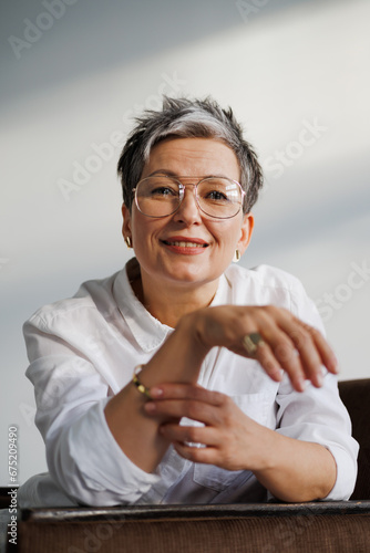 Smiling mature woman in white shirt and golden accessories looking at camera on grey 