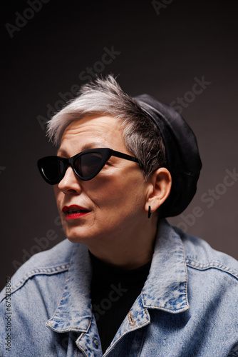 Portrait of fashionable mature woman in sunglasses, leather beret and denim jacket isolated on black  © Dmytro Hai