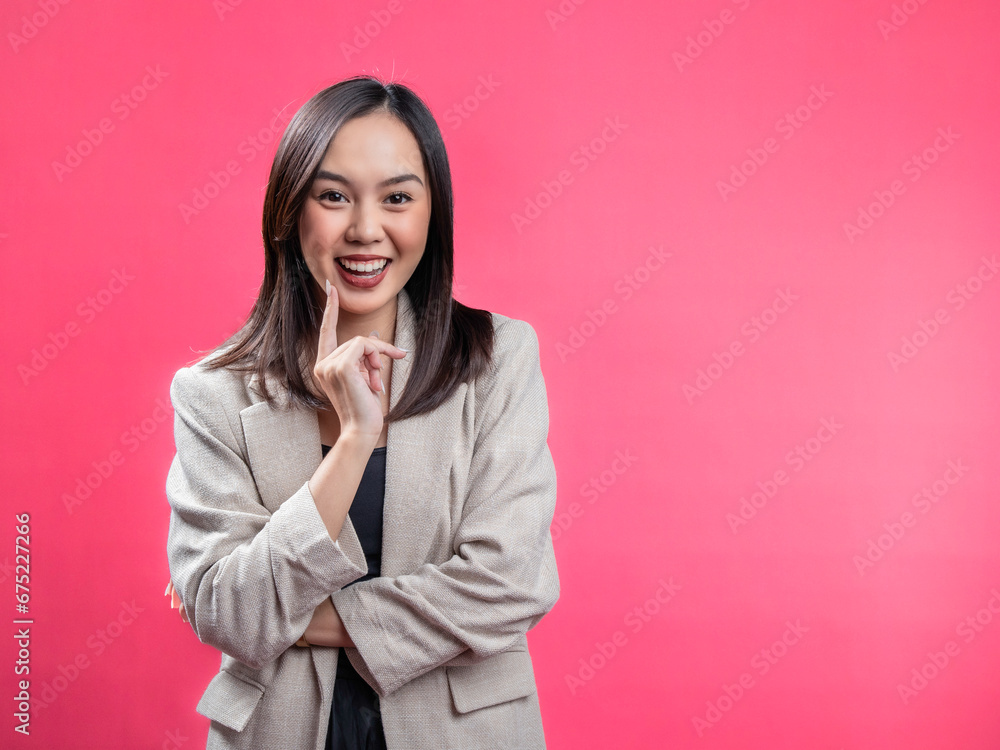 A portrait of an Indonesian Asian woman wearing a cream-colored blazer, posing happily with an index finger on her cheek. Isolated against a magenta background. A portrait of an Indonesian Asian woman wearing a cream-colored blazer, posing happily with an index finger on her cheek. Isolated against a magenta background.