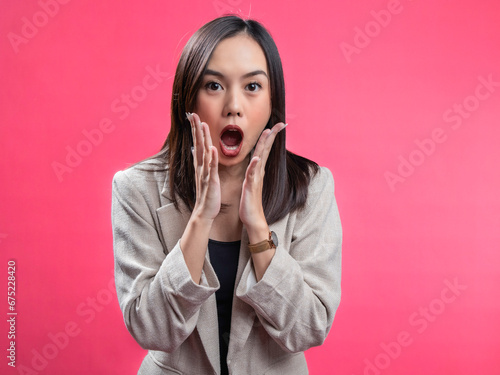 A portrait of an Indonesian Asian woman wearing a cream-colored blazer, looks surprised while opening her hands under her chin. Isolated against a magenta background.