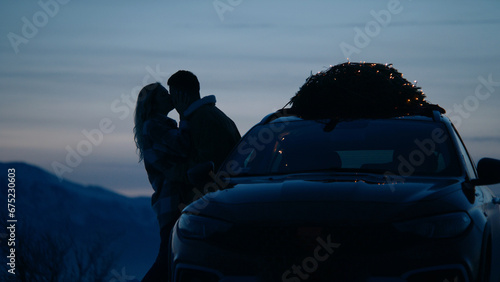 Silhouette of a couple kissing near the car with lit Christmas tree tied to the roof, winter mountains in the background (ID: 675230603)