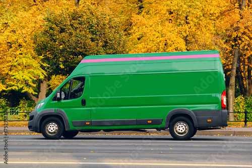 A green van drives along the road next to a city park in autumn. Fall has arrived, trees in colorful foliage.