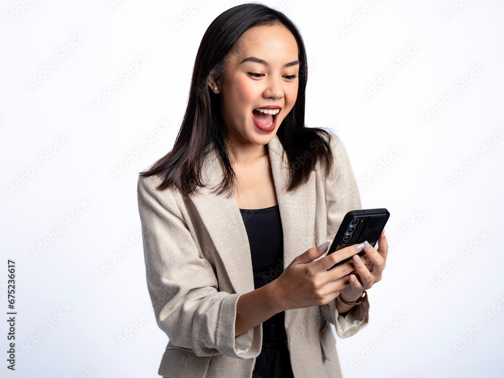 A portrait of an Indonesian Asian woman wearing a cream-colored blazer, looking happy while responding to a text message. Isolated against a white background. A portrait of an Indonesian Asian woman wearing a cream-colored blazer, looking happy while responding to a text message. Isolated against a white background.