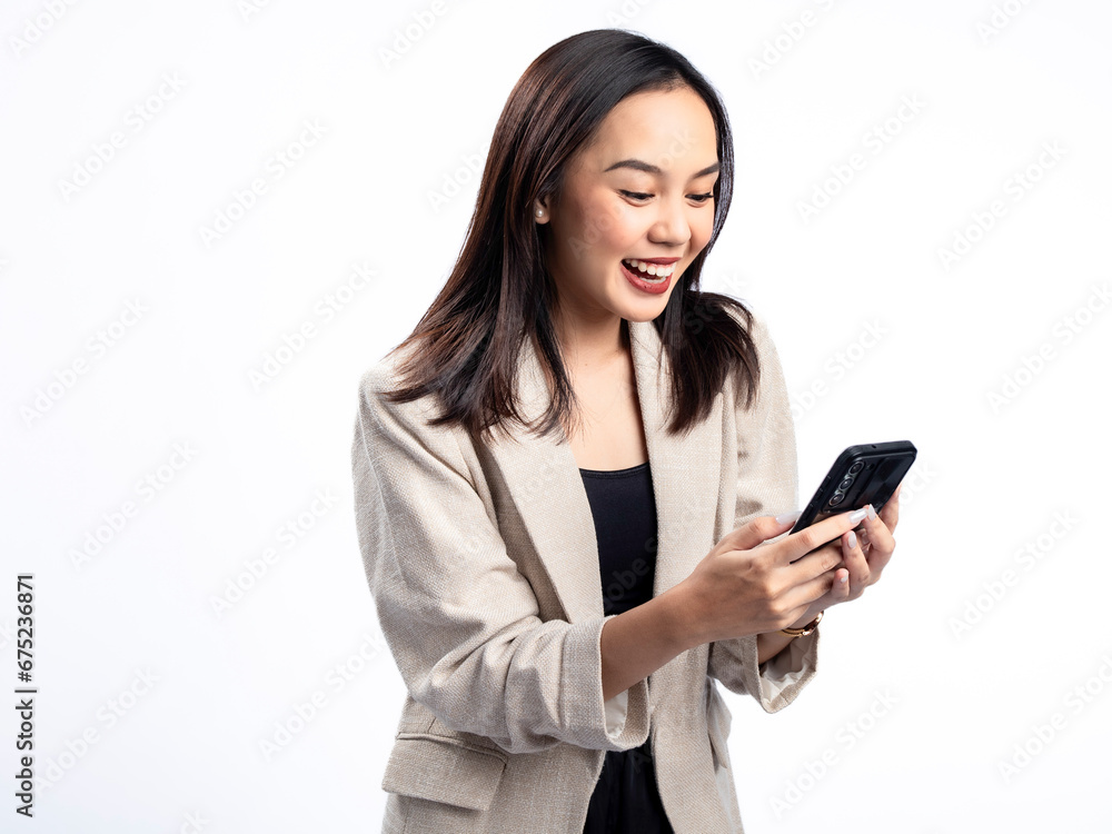 A portrait of an Indonesian Asian woman wearing a cream-colored blazer, looking happy while responding to a text message. Isolated against a white background. A portrait of an Indonesian Asian woman wearing a cream-colored blazer, looking happy while responding to a text message. Isolated against a white background.