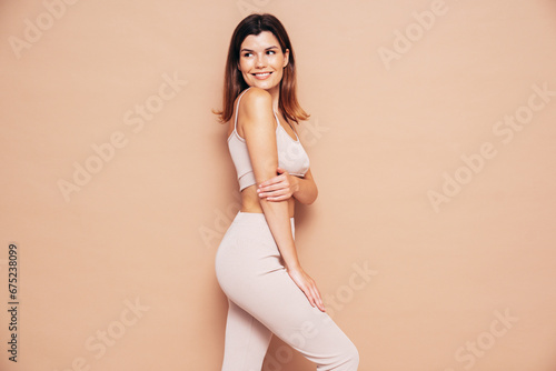 Young beautiful smiling female in trendy summer sport leggings and top clothes. Carefree woman posing near beige wall in studio. Positive model having fun indoors. Cheerful and happy