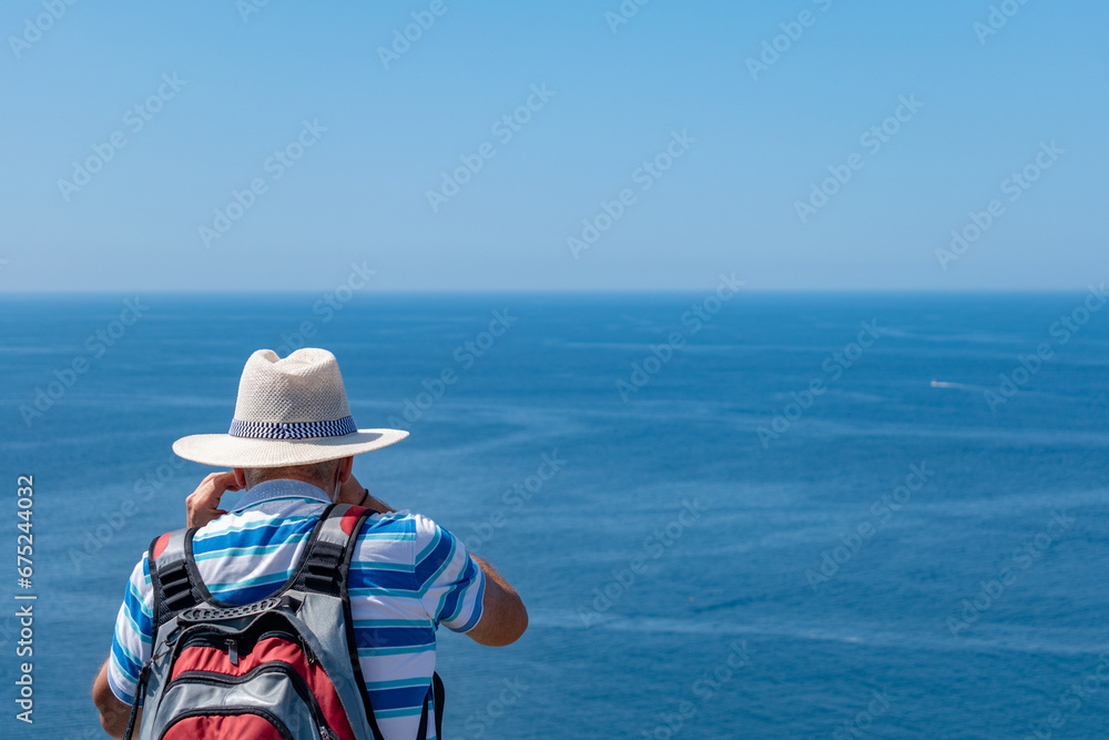 Senior man on his back with white sun hat and backpack photographing the Atlantic ocean. Tourism in summer. active seniors. Galicia, island of Ons. Copy space.