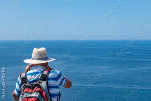 Senior man on his back with white sun hat and backpack photographing the Atlantic ocean. Tourism in summer. active seniors. Galicia, island of Ons. Copy space. photo