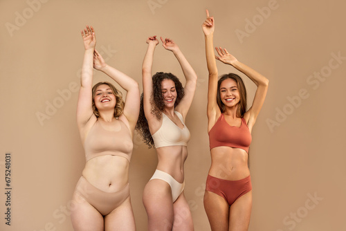 Self love concept. Cheerful ladies with hands up posing in only underwear over beige background. Caucasian women with different skin and body types expressing sincere support to each other in studio.