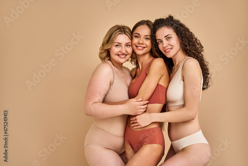 Natural female beauty. Group of three body positive women wearing comfy lingerie posing face to face and hugging in studio with beige background. Concept of body acceptance and support. Copy space.