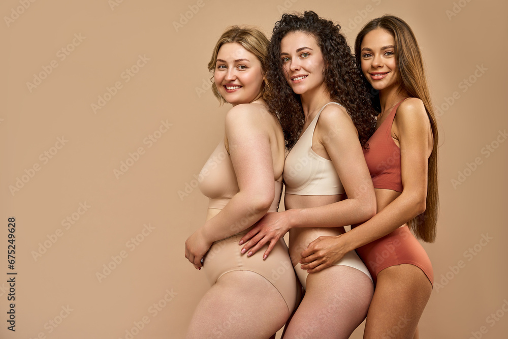 Love yourself. Stunning caucasian women in sporty beige, white and red bikini standing together over brown studio background. Three different ladies showing self love and body acceptance, copy space. - obrazy, fototapety, plakaty 