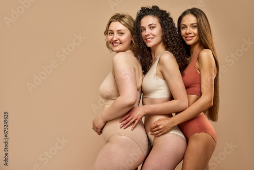 Love yourself. Stunning caucasian women in sporty beige, white and red bikini standing together over brown studio background. Three different ladies showing self love and body acceptance, copy space.