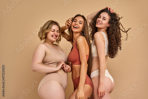 Happiness and self love. Group of three stunning women with beaming smile on faces and different body size posing together in tender lingerie in studio. Isolated over beige background. © Velista production