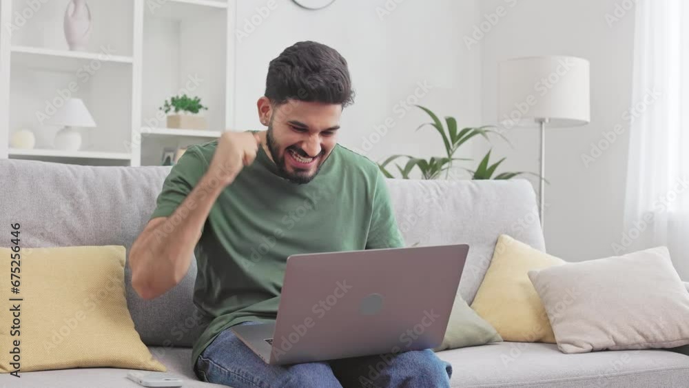 Happy hindu man wearing casual clothes sitting on couch with modern laptop and emotionally gesturing. Male freelancer rejoicing great news from work while staying at home. Happy hindu man wearing casual clothes sitting on couch with modern laptop and emotionally gesturing. Male freelancer rejoicing great news from work while staying at home.