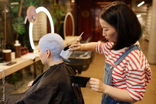 Side view portrait of young Asian woman as hairstylist applying bleach to short buzzcut hair in beauty salon
