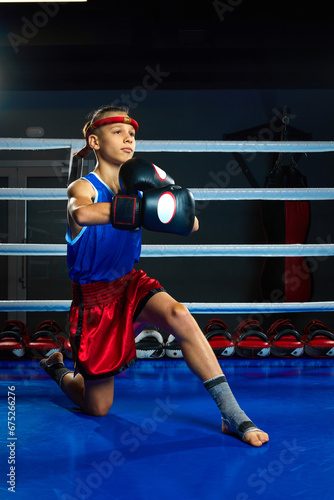 Serious boy, kickbox pupil, martial arts sportsman wearing uniform and gloves stands on knee and posing before fighting.